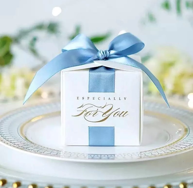 May include: A white gift box with a light blue ribbon and bow, sitting on a white plate with a gold rim. The box has a light blue vertical stripe and the words "Especially For You" in gold script.