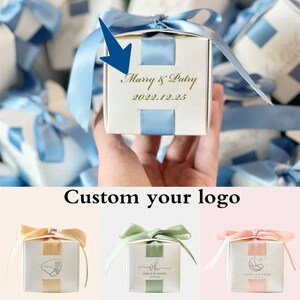 May include: A white gift box with a light blue ribbon and accents. The box is personalized with the text "Marry & Patry" and the date "2022.12.25". Additional boxes with different ribbon colors are shown.