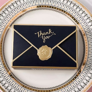 May include: A black envelope with a gold border and a gold wax seal. The envelope has the words "Thank You" written in gold script.