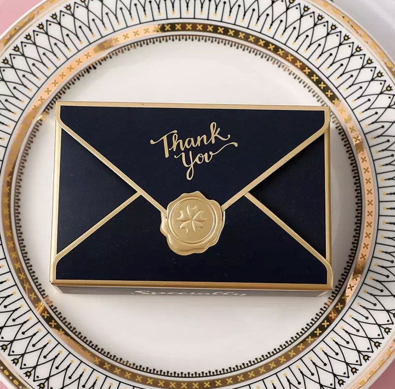 May include: A black envelope with a gold border and a gold wax seal. The envelope has the words "Thank You" written in gold script.