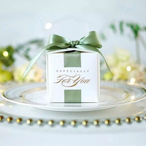 May include: A white gift box with a sage green ribbon and bow, sitting on a white plate with a gold beaded edge. The box has the words "ESPECIALLY For You" in gold script.