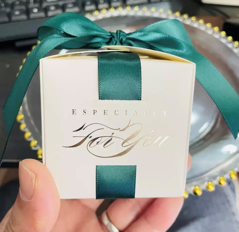 May include: A small, square gift box with a cream-colored exterior. The box is adorned with a dark green satin ribbon and bow. The words "ESPECIALLY For You" are printed in gold script on the front.