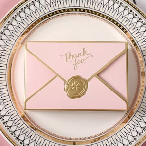 May include: Pink envelope with a gold border and a gold wax seal. The envelope has the words "Thank You" written in gold script.