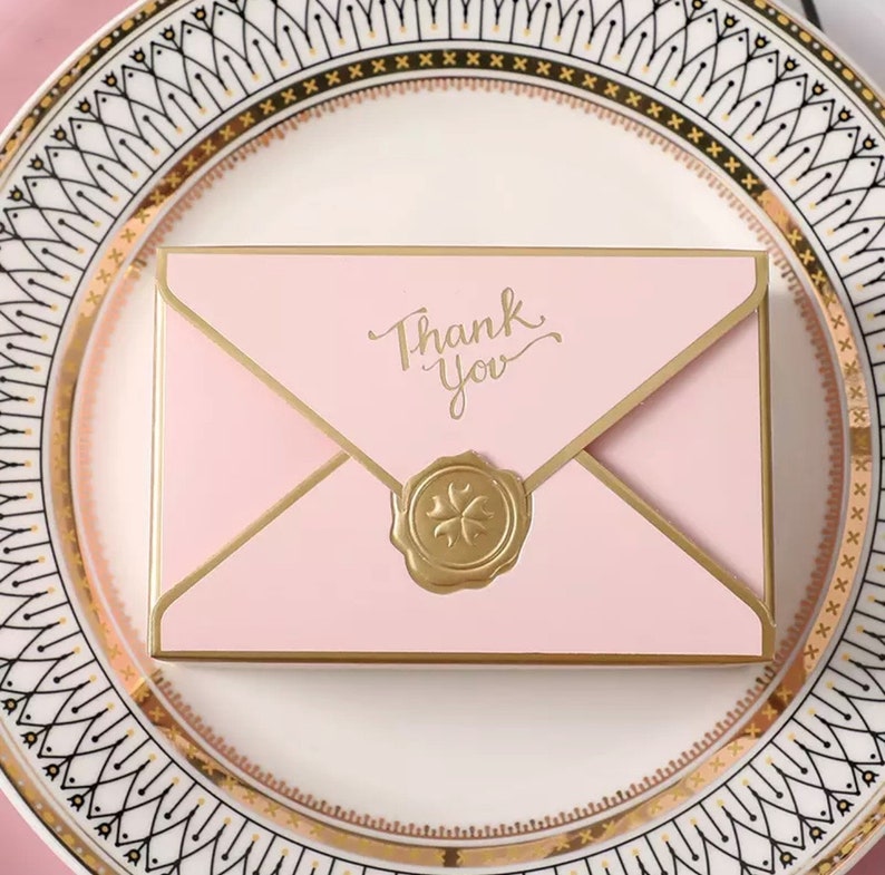 May include: Pink envelope with a gold border and a gold wax seal. The envelope has the words "Thank You" written in gold script.