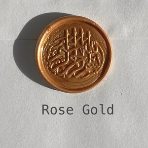 May include: A rose gold wax seal with an intricate design. The seal is round and has a raised design.