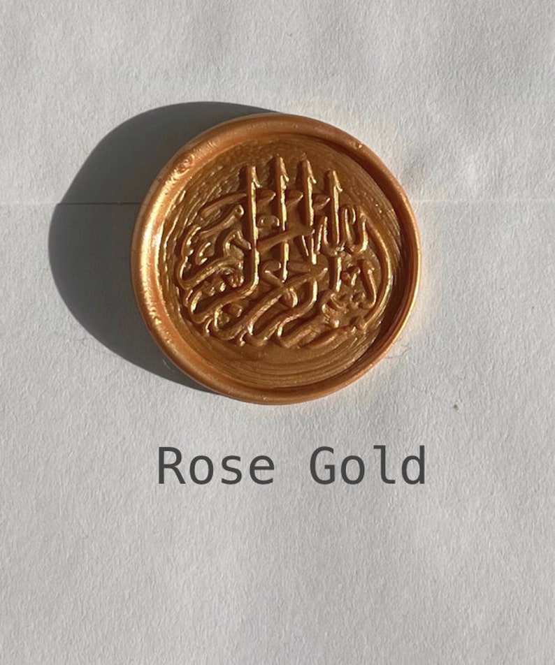 May include: A rose gold wax seal with an intricate design. The seal is round and has a raised design.