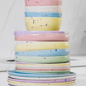 Könnte beinhalten: Ein vertikaler Stapel aus Keramikgeschirr, bestehend aus mehreren Schüsseln und Tellern in Pastelltönen wie Lavendel, Minzgrün, zartem Rosa, Himmelblau und Hellgelb, die alle ein mehrfarbiges Sprenkelmuster auf ihren glatten Oberflächen aufweisen.