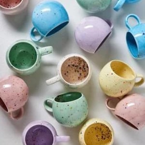 Op de afbeelding: Een verzameling gespikkelde keramische mokken in verschillende pastelkleuren, waaronder blauw, groen, roze, geel en paars. Elke mok heeft een afgeronde vorm met een klein handvat en een contrasterende binnenkleur. De mokken zijn gerangschikt op een wit oppervlak.