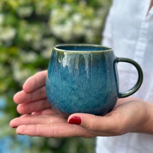 Könnte beinhalten: Eine blaue und grüne Keramiktasse mit abgerundeter Form und einem Henkel. Die Tasse hat eine gesprenkelte Glasur und eine leicht strukturierte Oberfläche.