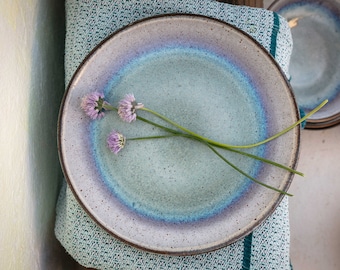May include: A ceramic plate with a speckled, light blue and purple glaze, with a dark brown rim. The plate is decorated with sprigs of purple flowers and green stems, resting on a teal cloth. Additional plates are stacked in the background.