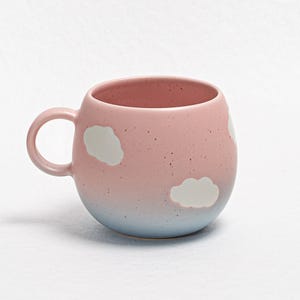 Puede incluir: Una taza de cerámica de color rosa y azul degradado con diseños de nubes blancas. La taza tiene una forma redondeada y un asa.