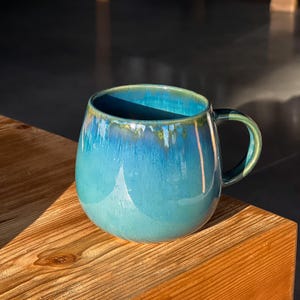Könnte beinhalten: Eine tealblaue Keramiktasse mit abgerundetem Körper und Henkel. Die Tasse hat eine einzigartige Glasur, die einen Marmoreffekt erzeugt.