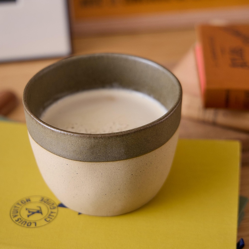 Op de afbeelding: Een keramische koffiekop met een lichtbeige buitenkant en een donkergroene rand, gevuld met een romige drank. De beker staat op een geel boek met tekst en een logo. De achtergrond omvat een houten oppervlak en een boek.