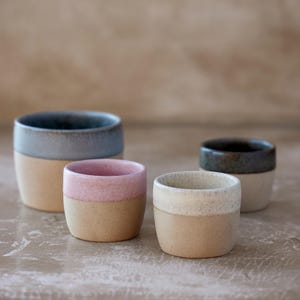 Puede incluir: Cuatro tazas de cerámica en varios tamaños y colores. Las tazas tienen una base beige natural con bordes de colores en azul, rosa y blanco roto. Las tazas están dispuestas sobre una superficie neutra, mostrando su diseño minimalista.