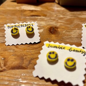 May include: Yellow smiley face stud earrings with purple smiles on white scalloped cards. The text "BUMBLE BEADSZ" is written in black on a yellow strip of paper above the earrings. The earrings are on a wooden surface.