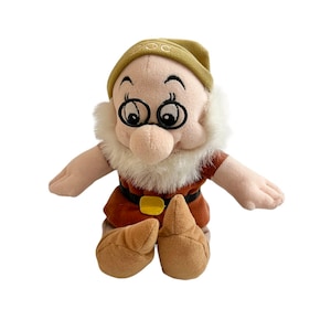 May include: A plush toy of Doc, one of the seven dwarfs from Snow White and the Seven Dwarfs. Doc is wearing a brown and tan outfit, a brown hat with the word "Doc" embroidered on it, and round glasses. He has a white beard and a friendly expression.