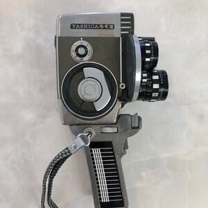 Peut inclure: Un appareil photo argentique Yashica 8-E III Super 8 vintage. L'appareil photo a un corps argenté et gris avec un objectif, des cadrans et une poignée avec une dragonne. L'appareil photo est sur une surface blanche.