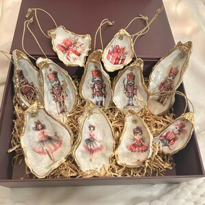 Op de afbeelding: Een verzameling oesterschelp-ornamenten met goudkleurige randen, elk met een uniek ontwerp. De ornamenten bevatten afbeeldingen van notenkrakers, ballerina's en ingepakte cadeaus, allemaal met gouden touwhangers, in een paarse doos.