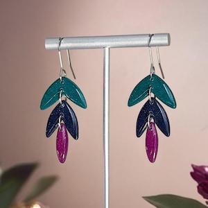 Può includere: Orecchini pendenti con un design a forma di foglia. Ogni orecchino presenta tre elementi a forma di goccia stratificati in verde acqua, blu navy e magenta. Gli orecchini sono esposti su un supporto argentato, con uno sfondo rosa tenue e accenti floreali.