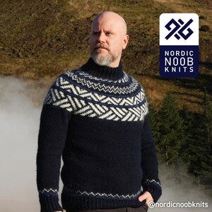 Könnte beinhalten: Ein marineblauer Pullover mit einem weißen und blauen geometrischen Muster. Der Pullover hat einen hohen Kragen und ist aus einem weichen, warmen Material gefertigt. Der Pullover wird von einer Person getragen, die vor einem malerischen Hintergrund steht. Der Text "NORDIC NOOB KNITS" ist in der oberen rechten Ecke des Bildes sichtbar.