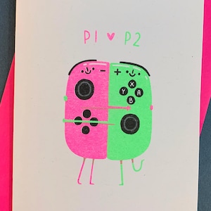 Op de afbeelding: Een wenskaart met een roze en groene illustratie van twee knuffelende gamecontrollers, met de tekst "P1 ♥ P2". De kaart ligt op een felroze envelop, tegen een donkerblauwe achtergrond.