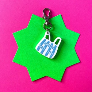 May include: A blue and white striped bag charm with the text "Bagsy Not It!" hanging from a silver key chain on a green star-shaped background.