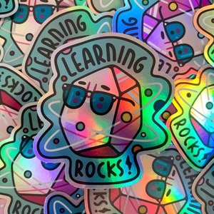 Puede incluir: Una pegatina holográfica con un cristal de dibujos animados que lleva gafas de sol y una burbuja de diálogo que dice "Learning Rocks!"