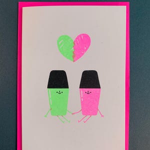 Op de afbeelding: Een wenskaart met een neonroze rand. De kaart toont een groen en roze hart boven twee stripfiguren die elkaars hand vasthouden. De figuren zijn groen en roze met zwarte tops en lachende gezichten.