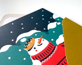 May include: A Christmas card featuring a snowman wearing a red and white sweater and a gold crown. The snowman is standing in a snowy landscape with green trees and white snow.