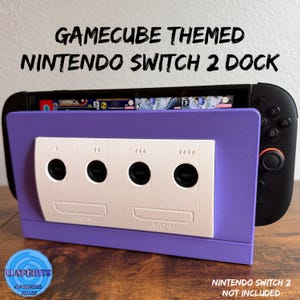 May include: A purple Nintendo Switch 2 dock with a GameCube theme. The dock features a white panel with four black circular ports and the text "GameCube Themed Nintendo Switch 2 Dock". The Nintendo Switch 2 is not included.