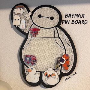 Puede incluir: Un tablero de pines con forma de Baymax, contorno negro e interior blanco texturizado. El tablero incluye varios pines de esmalte, como un robot rojo, un vehículo rojo, un pato y personajes. El texto dice BAYMAX PIN BOARD y BALALALALA.