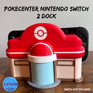 Op de afbeelding: Een rood en wit Pokecenter Nintendo Switch 2 Dock. Het dock is ontworpen om te lijken op een gebouw uit de Pokemon-serie. Het dock heeft een rood dak met een witte cirkel en een rood-witte koepel. Het dock is gemaakt van plastic.