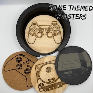 May include: A set of game-themed coasters. The coasters feature engraved designs of game controllers and consoles. The set includes a black coaster holder. The text "GAME THEMED COASTERS" is in the upper right corner.