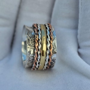 May include: A silver spinner ring with floral engravings and multiple bands of gold, copper, and silver. The ring is set against a white background, showcasing its intricate design and mixed metal elements. The ring's bands are twisted and smooth.
