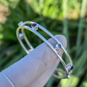 Peut inclure: Un bracelet jonc argenté à trois rangées, chacune ornée de petites pierres précieuses colorées. Les pierres comprennent des pierres rouges, bleues et transparentes. Le bracelet est présenté sur un fond vert flou.