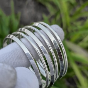 May include: A stack of shiny, silver bangles. The bracelets are smooth and reflective, with a simple, minimalist design. The bangles are stacked on top of each other, creating a layered effect. The background is blurred, with green and white colors.