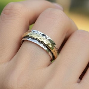 May include: A silver and gold-tone spinner ring with a textured hammered design. The ring has a rotating band. The ring is worn on a finger, showcasing its design and craftsmanship. The ring's design is suitable for everyday wear.