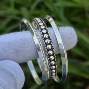Peut inclure: Trois bracelets en argent. L'un des bracelets présente une rangée de petites perles argentées en relief. Les deux autres bracelets sont lisses et ont un design simple et arrondi. Les bracelets sont présentés sur une surface blanche sur un fond vert flou.