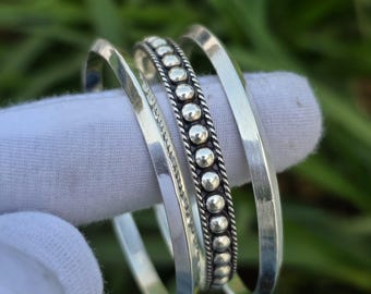 May include: Three silver bangles. One bangle features a row of small, raised silver beads. The other two bangles are smooth and have a simple, rounded design. The bangles are displayed on a white surface against a blurred green background.