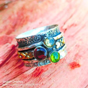 Może przedstawiać: Srebrny pierścień obrotowy z ozdobnym wzorem i czterema kamieniami szlachetnymi: przezroczystym, czerwonym, zielonym i złotym akcentem.