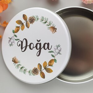 Puede incluir: Una lata redonda blanca con una tapa que presenta la palabra "Doğa" en una escritura elegante, rodeada por una corona de hojas, bellotas y bayas. La lata está abierta, revelando un interior metálico. Elementos florales decorativos en el fondo.