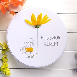 Puede incluir: Un objeto decorativo redondo blanco con una flor amarilla y una ilustración de dibujos animados de un niño con un gato. El texto "Hoşgeldin KEREM" está impreso en el objeto. Flores y follaje decorativos rodean el objeto.