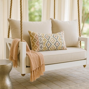 May include: A white wooden swing with beige cushions and a patterned throw pillow. The swing is hanging from a rope and is in front of a window with white curtains.