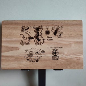 May include: A wooden cutting board with a map of the known world of Throne of Glass engraved on it. The map shows the continents of Crilea, Wendun, and the Southern Continent. The board is a light brown color and has a natural wood grain.
