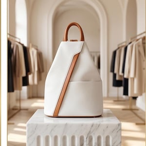 May include: A white backpack with a structured, triangular silhouette and a tan leather top handle. The bag features a diagonal tan leather accent strip running from the top to the bottom right corner, creating a clean, geometric design.