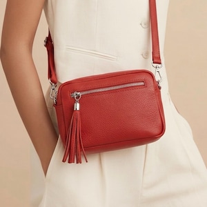 May include: A red leather crossbody bag with a silver zipper and tassel. The bag has a rectangular shape and a long, adjustable strap. The bag is approximately 9 inches wide and 6 inches tall.