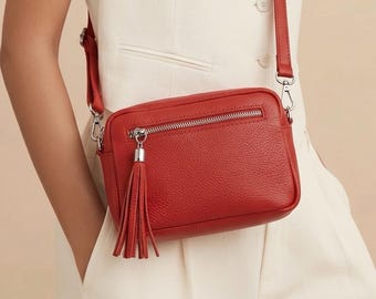 Puede incluir: Un bolso bandolera de cuero rojo con cremallera y borla plateadas. El bolso tiene forma rectangular y una correa larga y ajustable. El bolso mide aproximadamente 23 cm de ancho y 15 cm de alto.