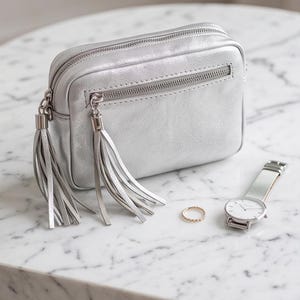 May include: A metallic silver rectangular crossbody bag with two long tassels attached to the zipper pulls, a thin gold-toned ring, and a silver-toned wristwatch with a white face and a mesh metal strap arranged together on a surface.