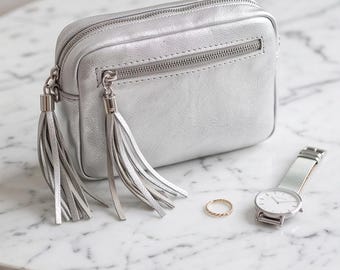 Puede incluir: Un bolso bandolera rectangular de color plata metalizado con dos borlas largas unidas a los tiradores de la cremallera, un anillo fino de tono dorado y un reloj de pulsera de tono plateado con esfera blanca y correa de malla metálica.