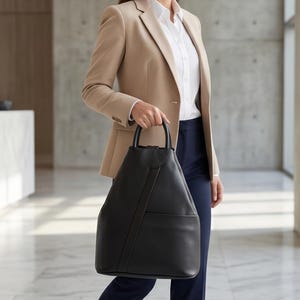 Może przedstawiać: Czarny sk&oacute;rzany plecak z g&oacute;rnym uchwytem i przednią kieszenią. Torba ma elegancki, nowoczesny design. Osoba ma na sobie beżową marynarkę, białą koszulę i granatowe spodnie.